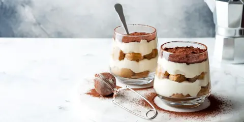 Dwa pucharki z tiramisu bez jajek, posypane kakao, na marmurowej desce.