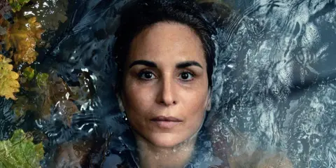 En kvinna med intensiv blick flyter i mörkt vatten, omgiven av höstlöv. En scen från veronica serie.
