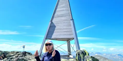En kvinna sitter på toppen efter att ha lyckats bestiga Helags. Hon ger tummen upp vid den ikoniska träpyramiden.
