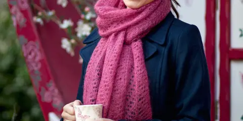 En kvinna ler och håller en mugg, insvept i en mjuk, rosa mohairhalsduk.