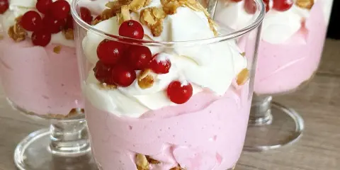 Różowy deser z galaretki z bitą śmietaną, chrupiącą granolą i czerwonymi porzeczkami w szklanych pucharkach.