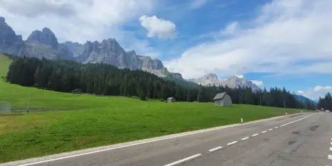 Strada panoramica verso il passo Monte Croce, con prati verdi, boschi di conifere e imponenti vette rocciose sotto un cielo azzurro con nuvole bianche.