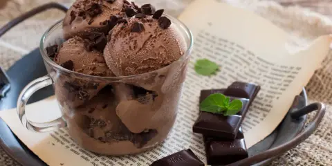 Coppa di gelato al cioccolato con scaglie di cioccolato fondente e foglioline di menta, su un vassoio arrugginito con un libro aperto.
