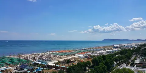Ampia spiaggia di Riccione con file ordinate di ombrelloni e lettini, bagnanti che si godono il sole e l'acqua cristallina del mare Adriatico sotto un cielo azzurro.