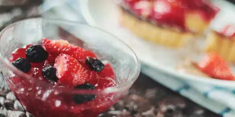 Szklana miseczka z galaretką truskawkową i owocami leśnymi, w tle deser z truskawkami.