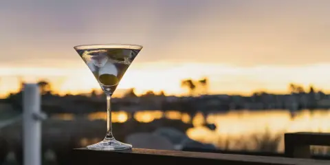 Idealny martini wstrząśnięte nie mieszane, z oliwką, na tle zachodzącego słońca nad wodą.