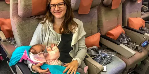 Uśmiechnięta kobieta trzyma roczne dziecko w samolocie. Otulone kocykami, maluch śpi spokojnie podczas podróży.