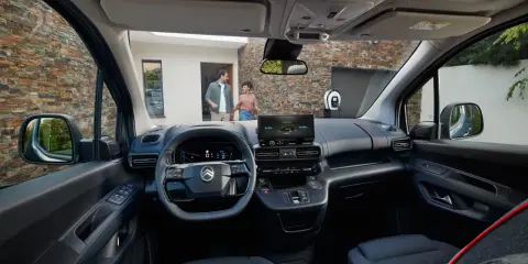 Interior de la nueva Citroën Berlingo, con pantalla táctil, volante moderno y asientos cómodos. Afuera, una pareja se acerca a un punto de carga.