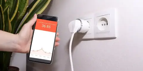 Smart wtyczka w gniazdku i smartfon z wykresem pokazującym monitoring zużycia energii.