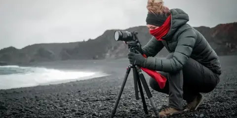 Fotografka na kamienistej plaży, używająca statywu, który przypomina monopody, by uchwycić surowe piękno krajobrazu.