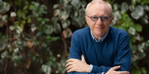David Grossman, con occhiali blu e maglione scuro, sorride con le braccia incrociate su sfondo verde.