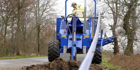 Máquina azul excava una zanja de drenaje, depositando tierra a un lado. Un operario supervisa el trabajo.