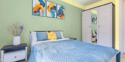 Dormitorio con paredes verde menta y detalles amarillos en el techo. Cama azul, mesita de noche y armario blanco. Inspiración de colores para pintar un dormitorio pequeño.