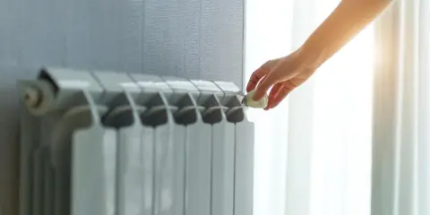 Dłoń reguluje termostat na grzejniku, pokazując różne rodzaje kaloryferów i kontrolę temperatury w domu.