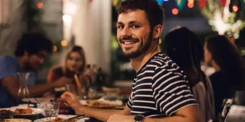Uśmiechnięty mężczyzna na tle przyjaciół przy stole. Idealny, tani pomysł na wieczór kawalerski z jedzeniem i dobrym towarzystwem.