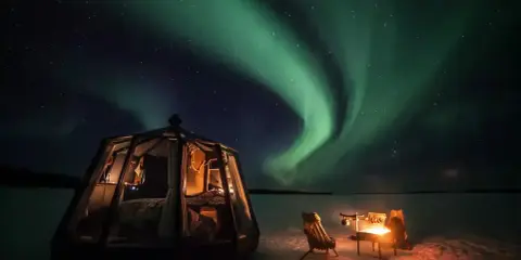 Drömmen att bo i igloo och se norrsken blir verklighet. En glaskupol med säng och en mysig eldstad utomhus under en magisk himmel.