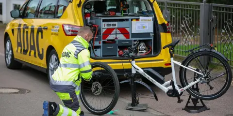 ADAC-Mitarbeiter repariert eine Fahrradpanne. Ein gelbes ADAC-Auto mit Werkzeugkasten steht daneben.