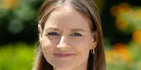 Porträt einer Frau mit blauen Augen und braunem Haar, das einige einzelne graue Haare abdeckt. Sie trägt goldene Ohrringe.