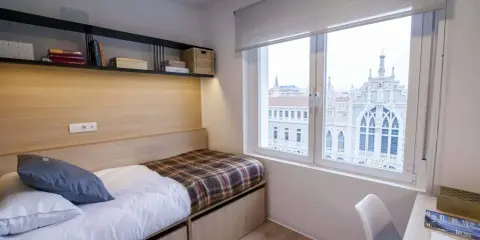 Habitación moderna con cama, estanterías y vistas a un edificio histórico en Xior Madrid Retiro.