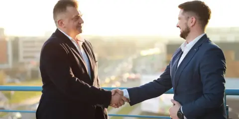 Dwóch mężczyzn w garniturach ściska sobie dłonie na tle miasta.