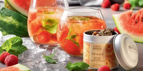 Rafraîchissante recette thé glacé : deux verres remplis de glaçons, fruits rouges et menthe, accompagnés de pastèque et d'une boîte de thé.