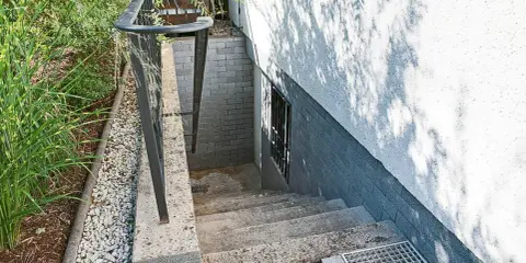 Betontreppe sanieren: Alte Steintreppe mit Geländer führt zu einer Kellertür. Grüne Pflanzen und Kies säumen den Weg.