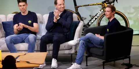 Drei Männer, darunter ein Fahrer von Jumbo-Visma, sitzen auf Sofas vor einem Rennrad.