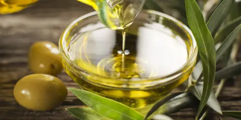 Olio di oliva non extravergine versato in una ciotola di vetro, con olive e foglie d'ulivo su fondo di legno.