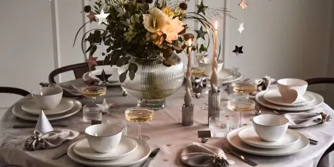Enkel nyårsdukning med vita tallrikar, champagneglas, stjärnformade dekorationer och en blomsterbukett i en glaskaraff.