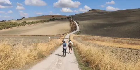 Due pellegrini camminano su una strada sterrata in un paesaggio collinare, forse da dove parte la Via Francigena.