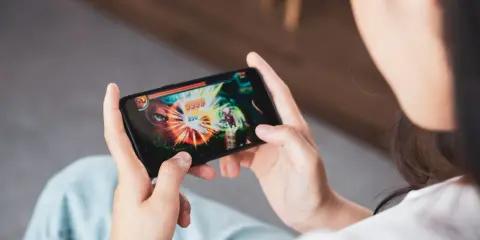 Osoba gra w gry dla dwóch osób na odległość na telefonie. Na ekranie widać dynamiczną scenę walki z potężnym przeciwnikiem.