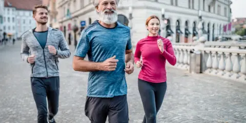Un uomo con barba bianca e due giovani corrono sorridenti in città. Un esempio di come iniziare a correre a 50 anni sia possibile e gratificante.