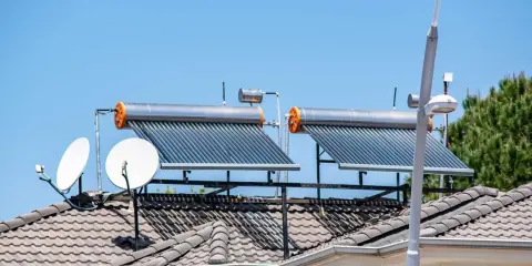 Dos calentadores de agua caliente solar en un tejado, junto a antenas parabólicas, bajo un cielo azul.
