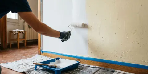 Persona aplicando pintura blanca con rodillo en pared. Se muestra bandeja con pintura y brocha. Aprende como hacer gotele con rodillo.