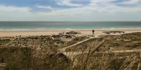 Strand von Conil de la Frontera mit Dünen, einem Strandcafé und Sonnenschirmen. Das Meer ist ruhig, der Himmel ist bewölkt.