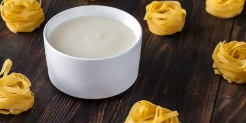 Un bol de béchamel sans beurre crémeuse, entouré de nids de pâtes fraîches sur fond de bois sombre.