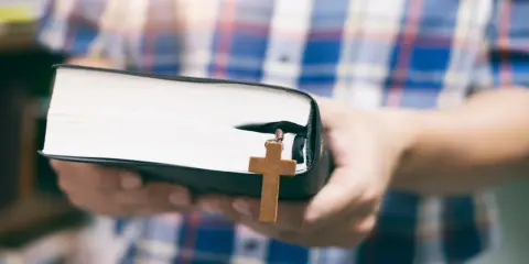 Osoba w kratę trzyma Biblię z drewnianym krzyżem, symbolizującym dwa przykazania miłości.