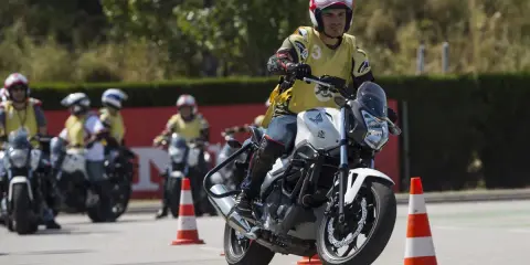 Hombre en moto blanca, con chaleco amarillo y casco, practicando maniobras en un curso carnet A. Otros motoristas y conos en el circuito.