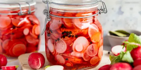 Zwei Gläser mit eingelegten Radieschen, leuchtend rot und mit schwarzen Pfefferkörnern. Frische Radieschen liegen daneben.