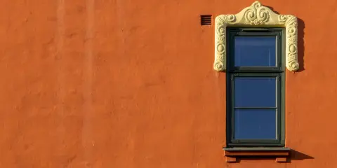Ett fönster med en utsmyckad, gul ram på en orange, putsad fasad. Man undrar om det är här man måste måla putsad fasad för att bevara dess charm.