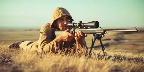 Mężczyzna w kapturze leży na trawie, celując z karabinu z lunetą.