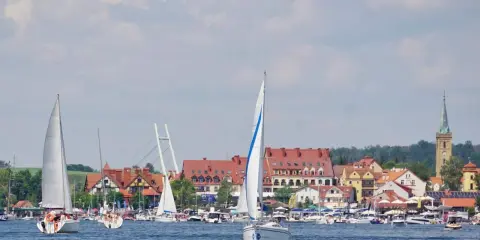 Żaglówki na jeziorze przed malowniczymi mazury miasta z czerwonymi dachami i wieżą kościelną.