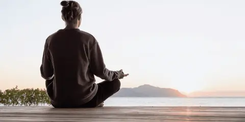 Person sitter i lotusställning på en träbrygga vid havet, med solnedgång i bakgrunden. En stund av mindful yoga.