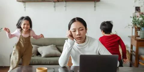 Donna esausta davanti al laptop, mentre i bambini giocano. Un quadro di **parental burnout** in casa.