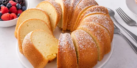 Pokrojona babka piaskowa, oprószona cukrem pudrem, z miseczką owoców leśnych w tle.