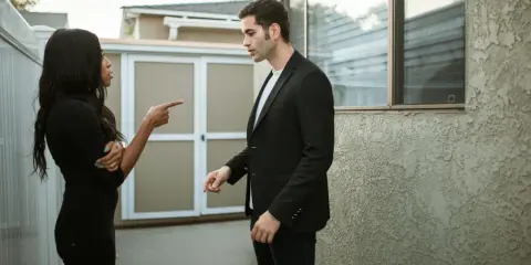 Une femme noire en robe noire pointe du doigt un homme en costume noir, lui reprochant quelque chose. Elle semble vouloir lui faire regretter quelqu'un.
