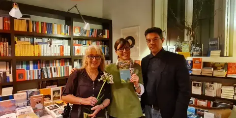 Tre persone sorridono in una libreria, una tiene in mano il libro "Cocco e Magella". Libri in ordine cronologico sugli scaffali.
