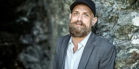 Man med skägg och keps, i grå kavaj, står mot en stenvägg. Kanske en del av en förintelsen serie på SVT.