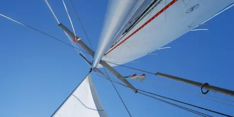 Vue plongeante sur le mat de bateau, voiles blanches déployées sous un ciel bleu éclatant, cordages tendus.