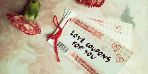 Papierowa rocznica uczczona kuponami miłości. Dwa kwiaty goździka i stosik kuponów "Love Coupons for You" na różowym tle.